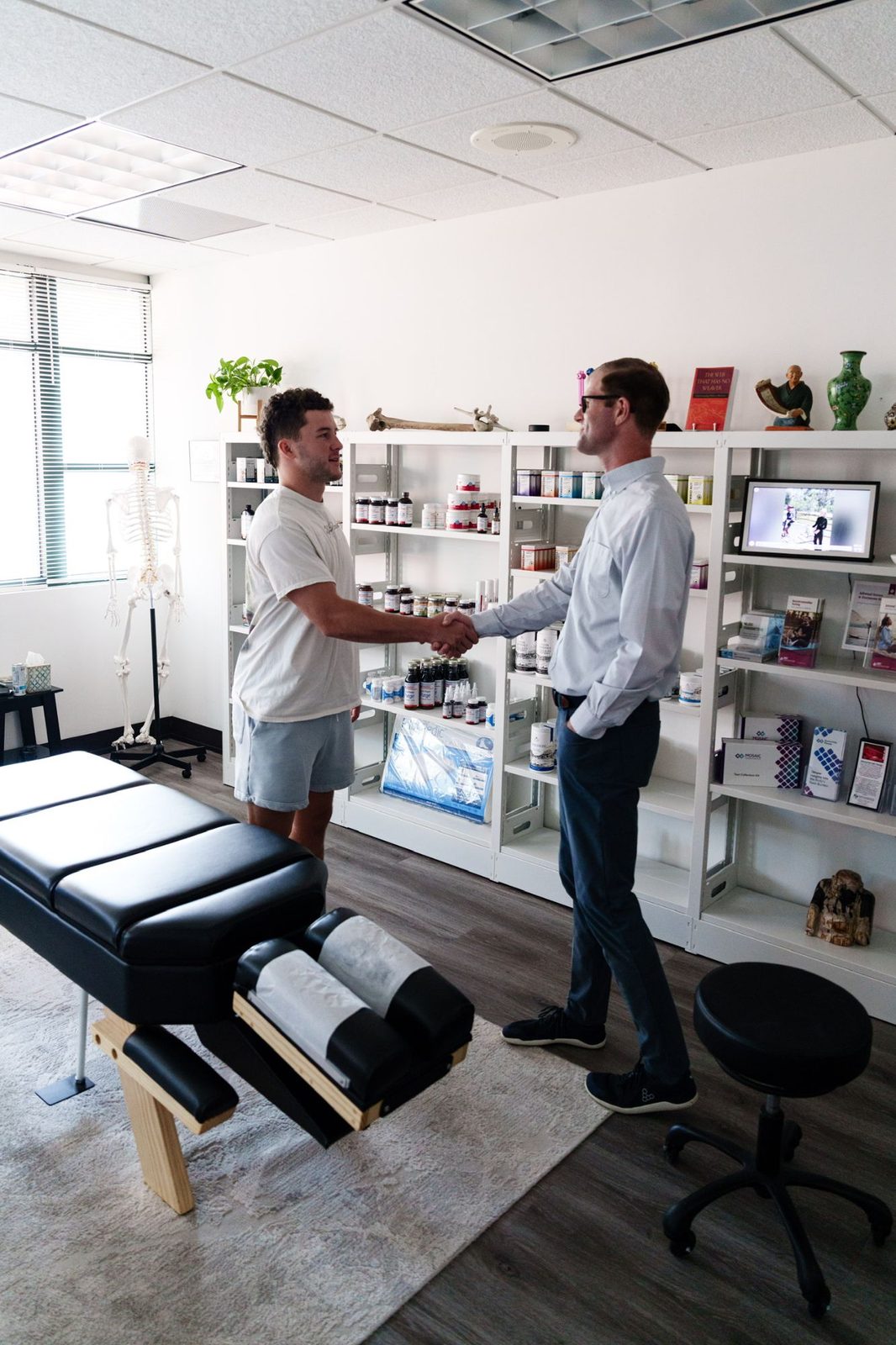 Dr. Sean Olenek greeting a visitor in the treatment room, with the supplement shelves and adjustment table